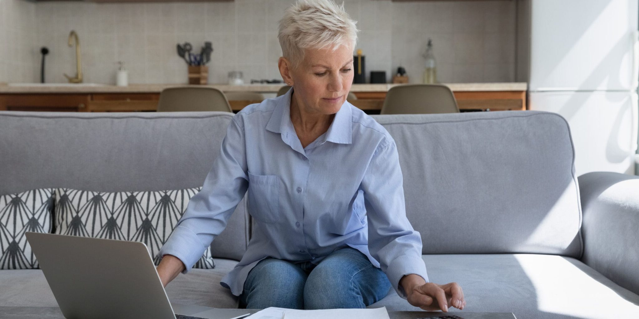 Serious attractive mature woman sit on sofa calculates expenses on calculator, use laptop paying bills through e-bank app at home. Family budget control, financial management, accounting job at home